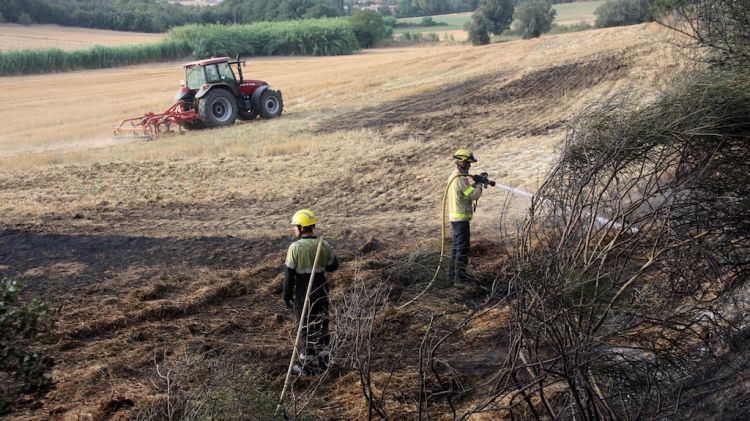 Bombers apagant l'incendi en un camp amb un tractor al fons llaurant © ACN