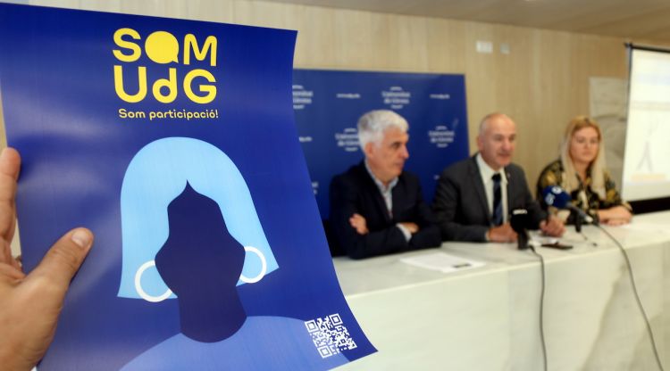 En primer terme, el cartell anunciant la plataforma 'Som UdG'; al fons, el rector Quim Salvi i el vicerector José Antonio Donaire. ACN