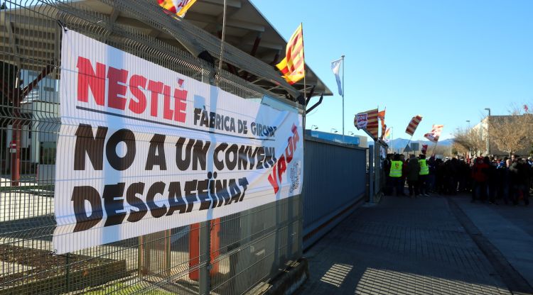 Una pancarta a favor de la vaga; al fons, els treballadors de la Nestlé de Girona concentrats. ACN