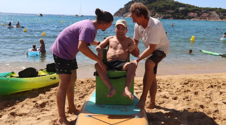El director de l'escola nàutica de la Fosca i un monitor de l'Associació Pots ajuden a una persona amb mobilitat reduïda a pujar a una planxa de paddle surf adaptat. ACN