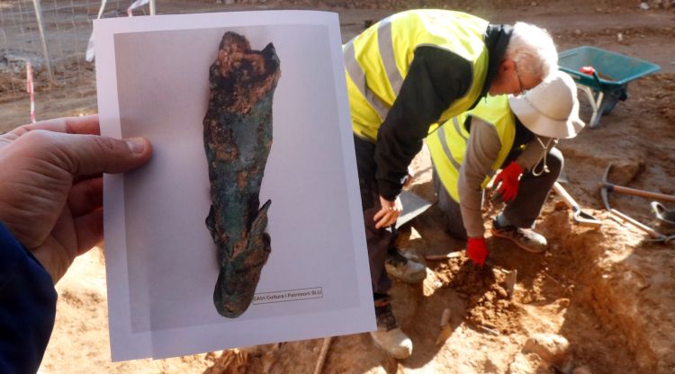 En primer terme, el fragment d'estàtua del déu Mercuri; al fons, els arqueòlegs excavant la necròpolis medieval. ACN