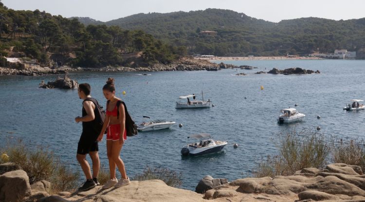 Dos joves passant pel camí de ronda que passa per davant d'un camp de boies amb embarcacions a Palamós. ACN