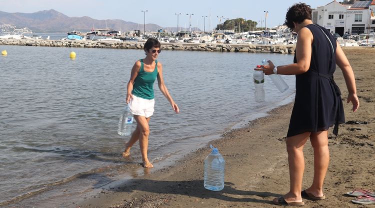 Dues dones omplen ampolles i garrafes del mar a la platja del Port de la Selva. ACN